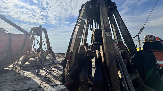 A close-up photo of a grapple which attaches to a crane. The grapple is resting on a barge in the water. The grapple has six arms and a pully system in the center. Another grapple is in the background of the photo.